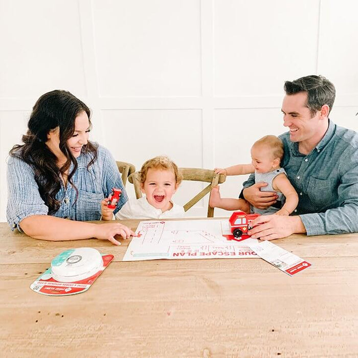 A man and woman with their two infant children sit at a table with First Alert safety products and a map of an escape route in case of a fire at home.
